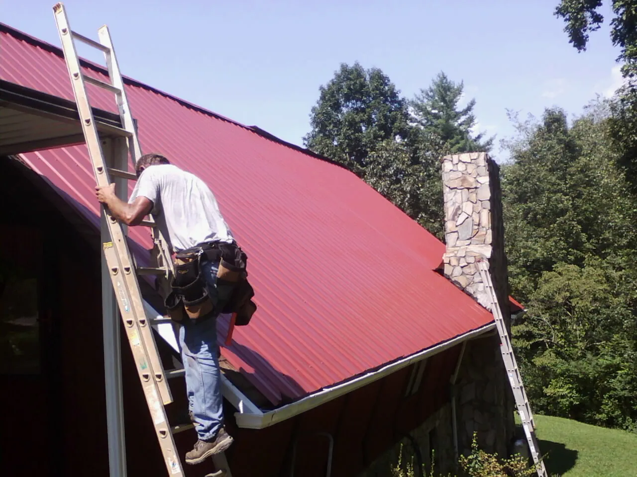 Roofer on a ladder beside a red metal roof and stone chimney — Soffit & Fascia Repair by Roofing Brothers in Husum